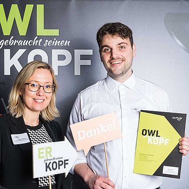 2 Personen stehen vor einer Wand mit dem Motiv "Stiftung Studienfonds OWL".